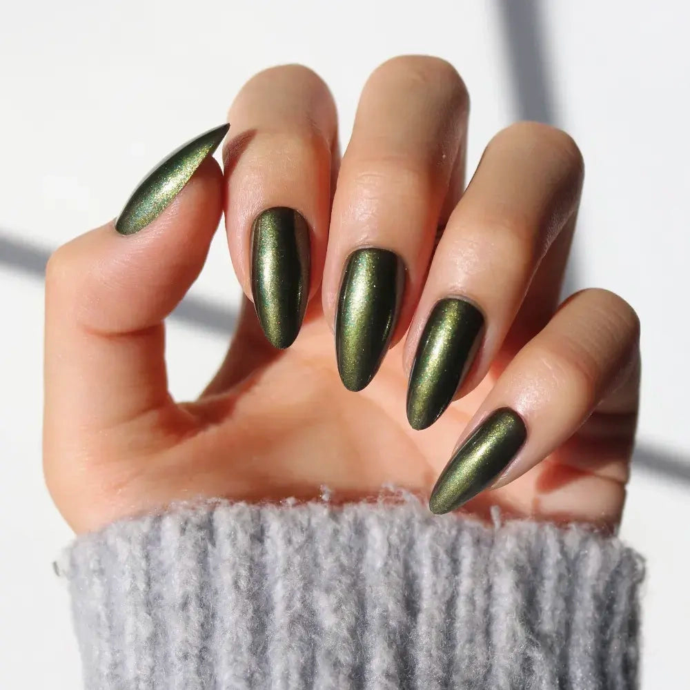 A hand with a glamnetic Cutie Pine metallic green almond nail set held up against a light background. The person is wearing a gray ribbed sweater.