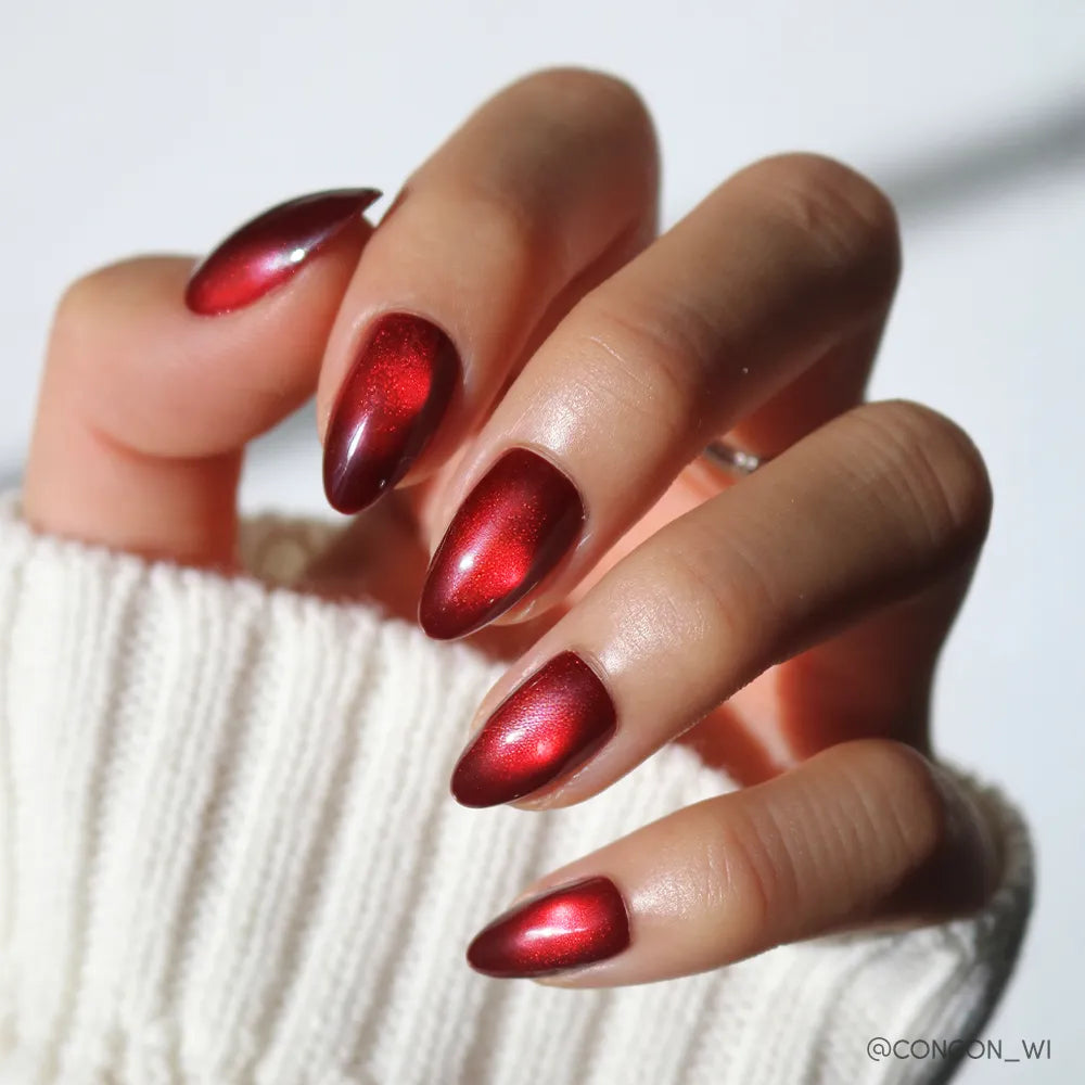 A hand with short almond nails, painted in glossy metallic red from glamnetic’s Frosted Cranberry set, is seen against a white knit sweater background.
