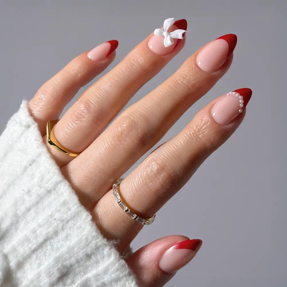 A hand with short almond nails featuring glamnetic’s Ruby Ribbons—red French tip press-ons with a 3D white flower accent on the index finger, pearls on the ring finger, and gold rings—styled with a cozy white sweater for the perfect festive holiday manicure or to show off your favorite glamnetic nail set.