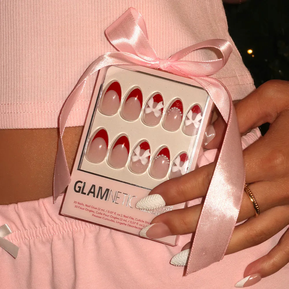 A hand with long white textured almond nails holds a glamnetic Ruby Ribbons press-on nail box with red French tips, ruby ribbons, and floral designs tied with a pink ribbon. The person wears pink and a gold ring—perfect for a holiday manicure. - Product image