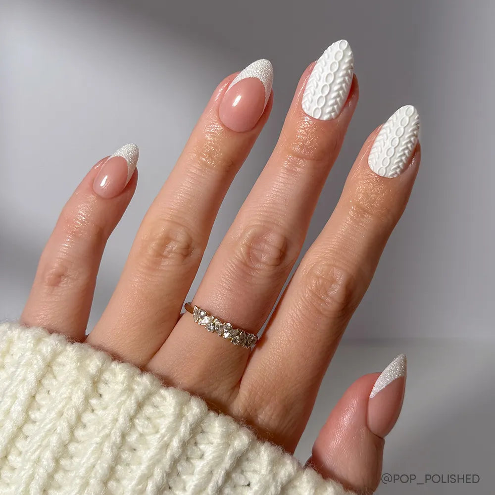 A hand with short almond nails features glamnetic’s Sweater Weather set: white textured sweater patterns and glitter French tips. The wearer is dressed in a knit sweater and sports a delicate ring with small stones.