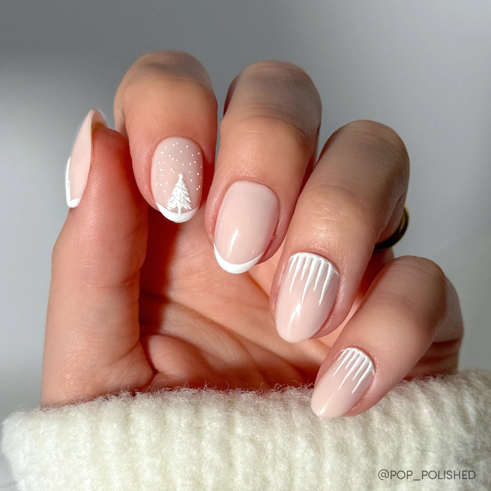 A hand wears glamnetic&#39;s Winter&#39;s Tale: short, oval Christmas nails with white French tips, a snowy tree with dots, and icicle-like vertical lines, finished with a cozy white sweater for a festive winter look.