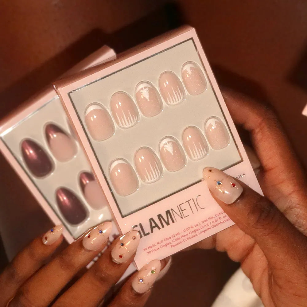 A person holds two press-on nail boxes—one with glossy pink nails, and one from glamnetic called Winter’s Tale, featuring nude winter nails with white flame designs. Their own nude nails have colorful star decals for a chic Christmas look. - Product image