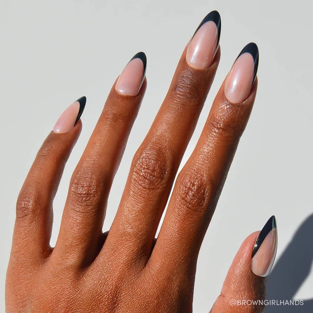 A hand with medium brown skin shows off glamnetic&#39;s Black Truffle nails: long, almond-shaped, in a nude shade with sleek black French tips. The background is bright and plain.