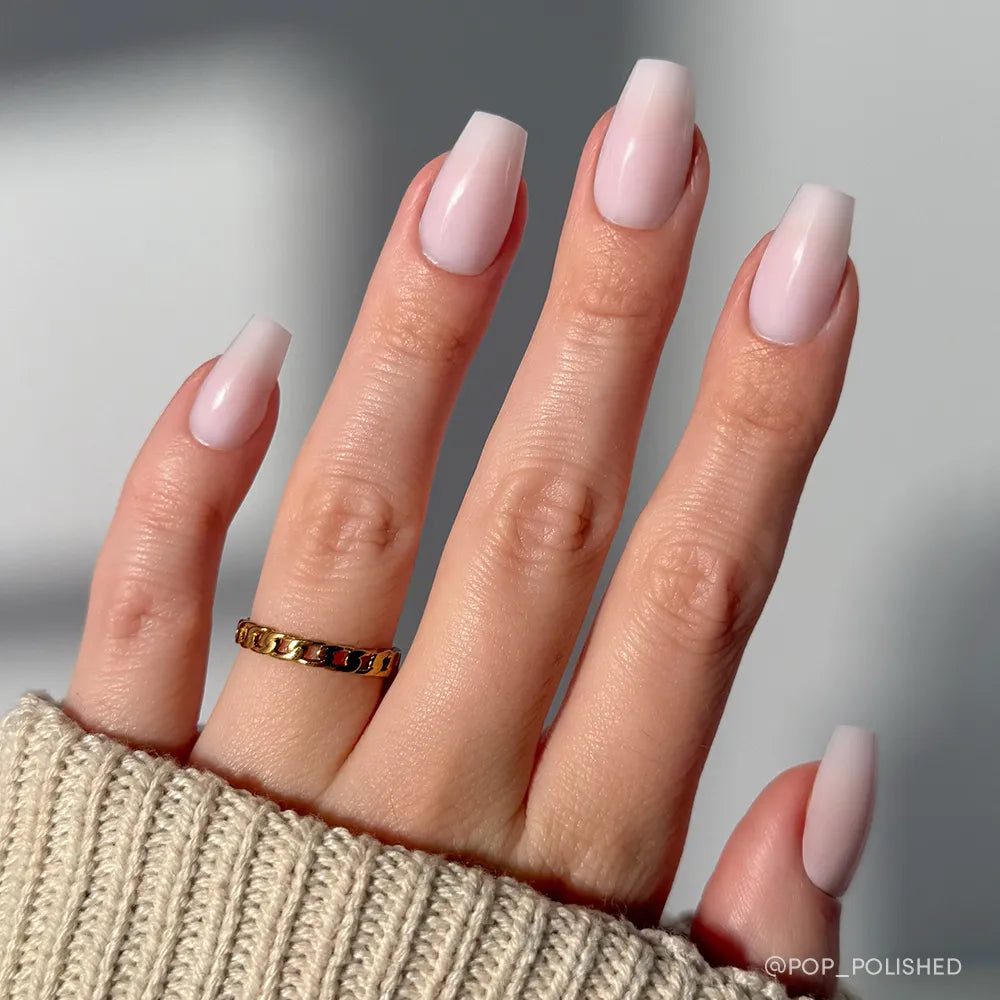 A hand with long, medium coffin nails in Blush Dream by glamnetic—a pale soft pink shade. The person wears a gold ring and beige knitted sweater. The background is blurred.