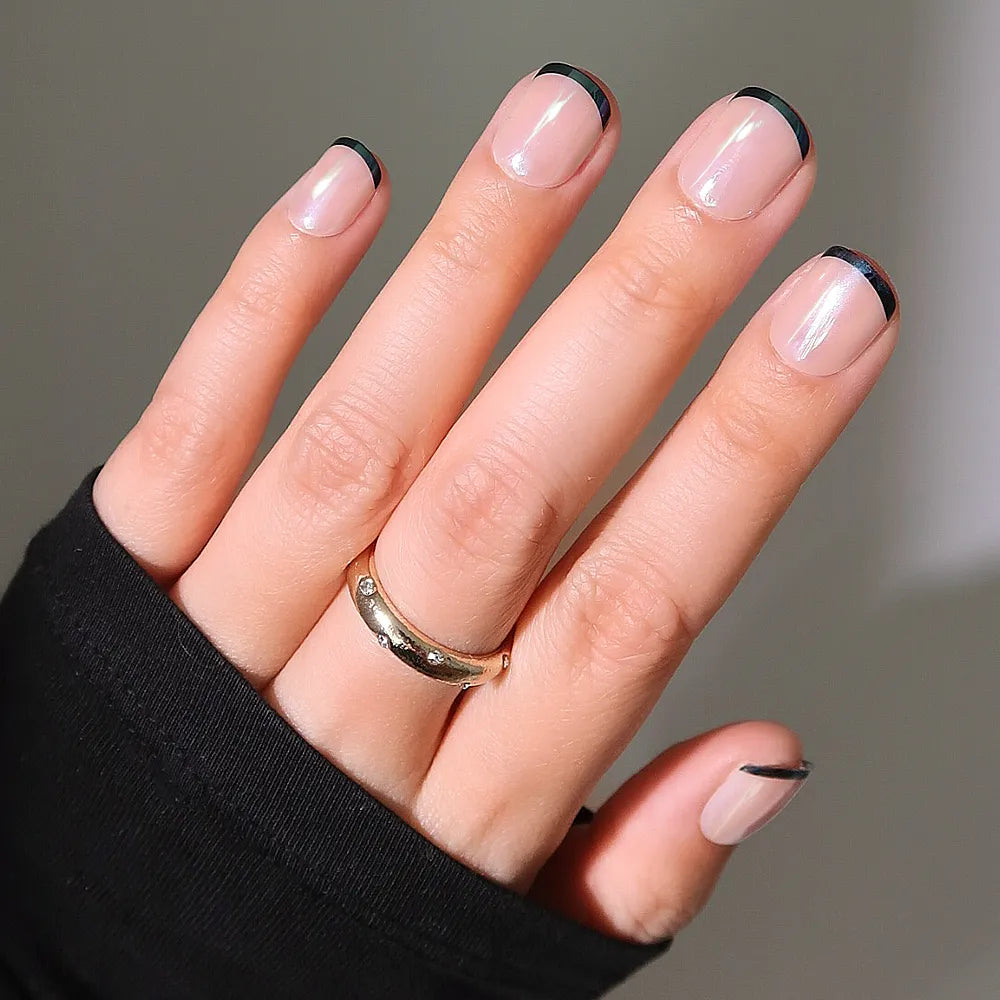 A hand with short, natural nails wears glamnetic&#39;s Burnt Miso, a minimalist French manicure with thin black tips and a glossy finish. The person sports a gold ring and a black long-sleeve top.