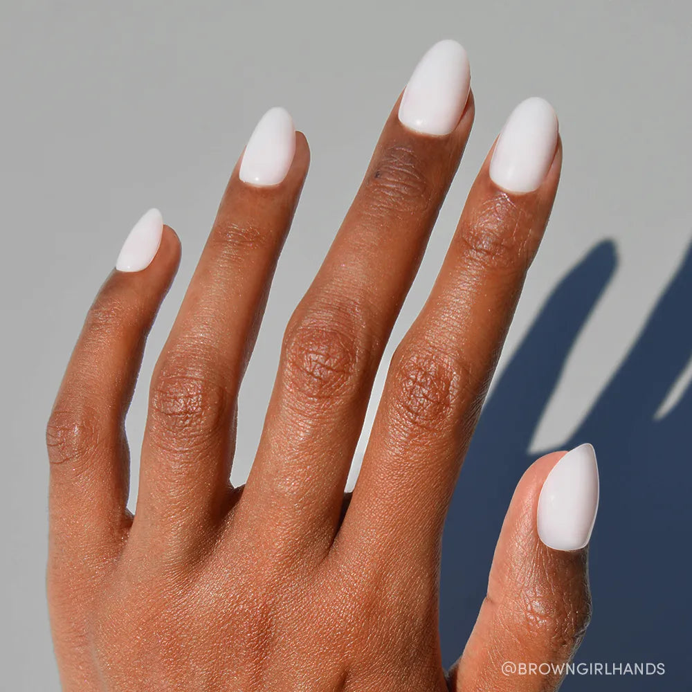 A hand with medium brown skin and short oval nails wears glamnetic&#39;s Clean Girl milky manicure, displayed against a neutral background with visible shadow.