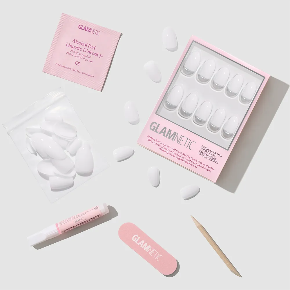 The glamnetic Clean Girl set includes milky white press-on nails, a pink nail file, a wooden cuticle stick, glue, an alcohol pad, and an extra pouch of short oval nails arranged neatly on a light gray surface. - Product image
