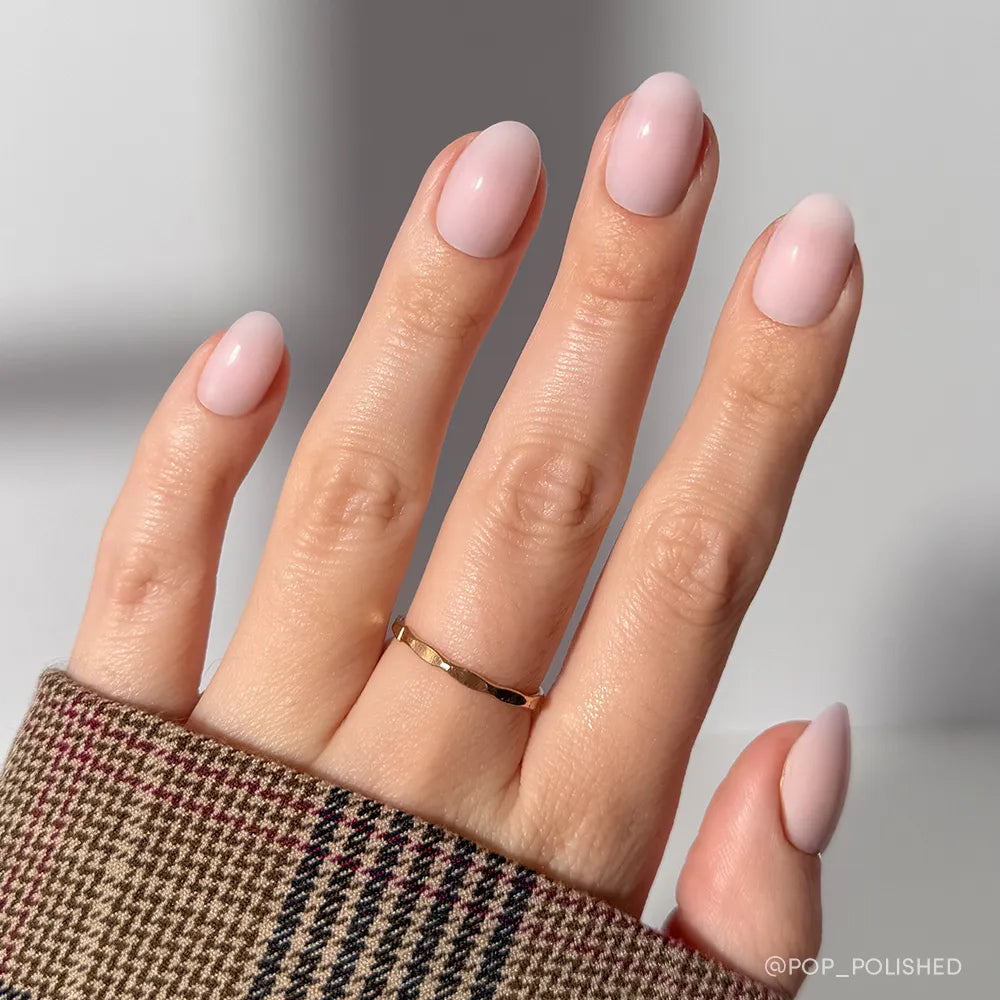 A hand with short oval nails painted in glamnetic’s Doll Cheeks—a light pink, salon-quality manicure. The person wears a thin gold ring and a brown plaid sleeve. The softly blurred background features neutral tones.