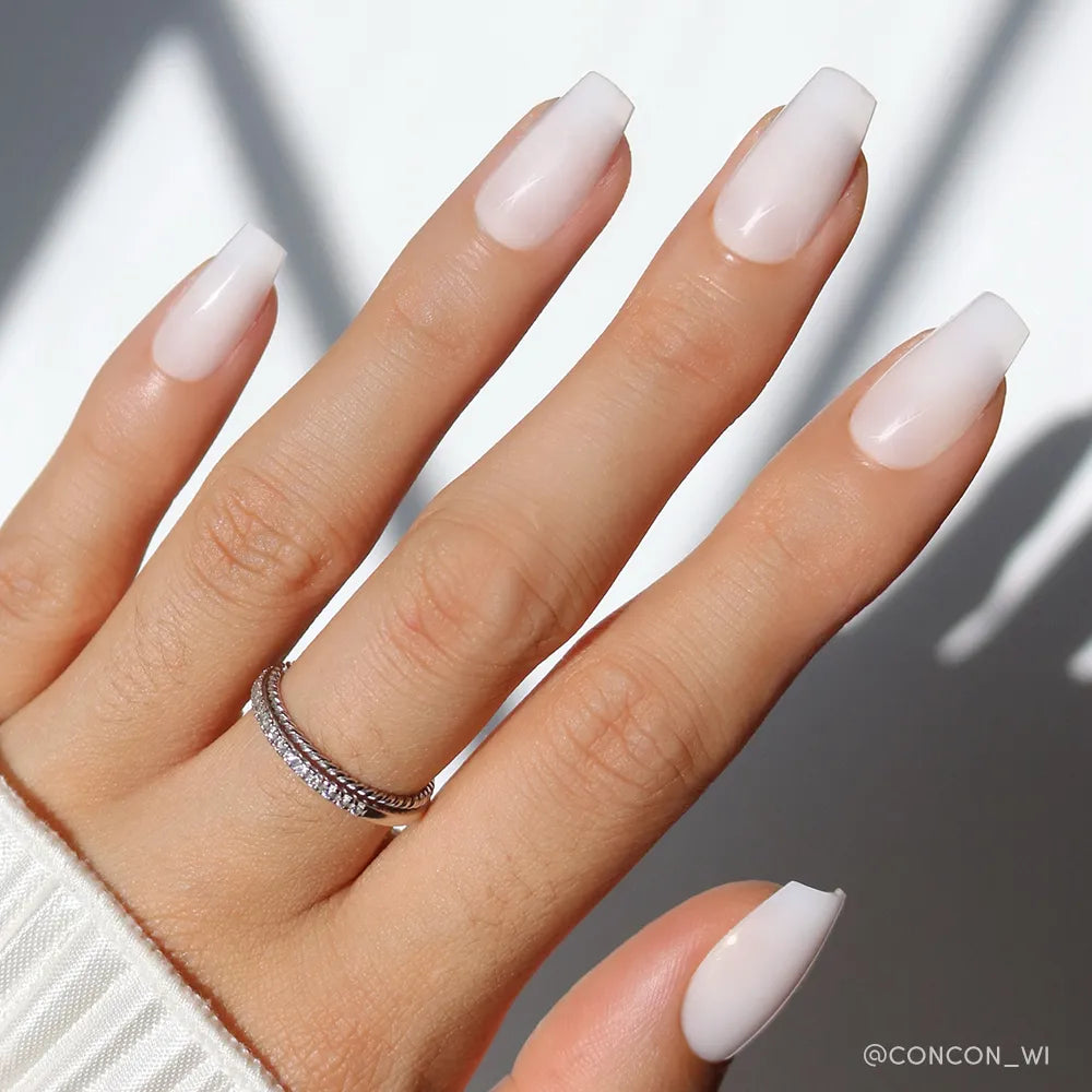 A hand with long, coffin-shaped nails features glamnetic&#39;s Oat Milk polish—a glossy, pale pink ombre for a chic milky mani. Two thin silver rings adorn the ring finger against a softly lit background with gentle shadows.