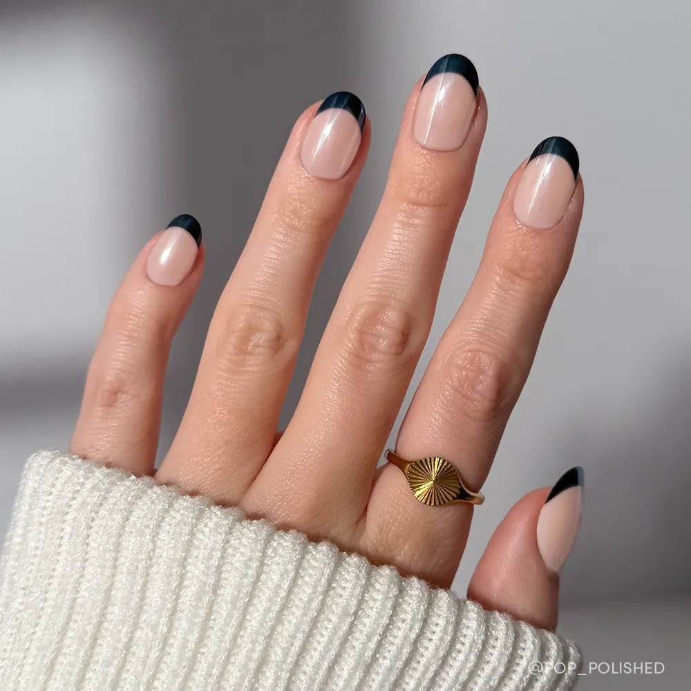 A hand showcases glamnetic&#39;s Onyx Glaze press-on nails with minimalist black French tips and a glossy finish. A gold sunburst ring and a white ribbed sweater sleeve add an elegant touch to the look.
