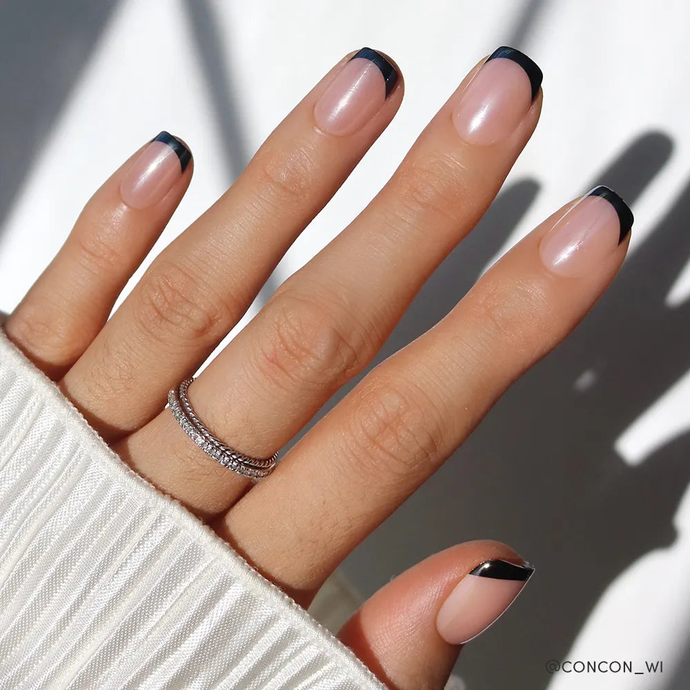 A hand with a light skin tone features glamnetic’s Squid Ink minimalist black French tip nails with a glossy finish, accessorized by two thin sparkly silver rings and a white pleated sleeve. - Product image