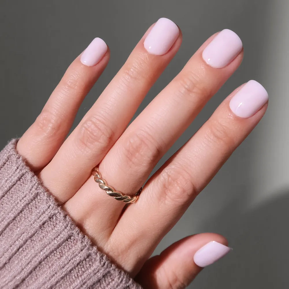 A hand with super short, light pink nails from the glamnetic Sweetie Set is shown. The person wears a gold twisted ring on their ring finger, with a mauve knit sweater sleeve partially covering their salon-quality manicure.