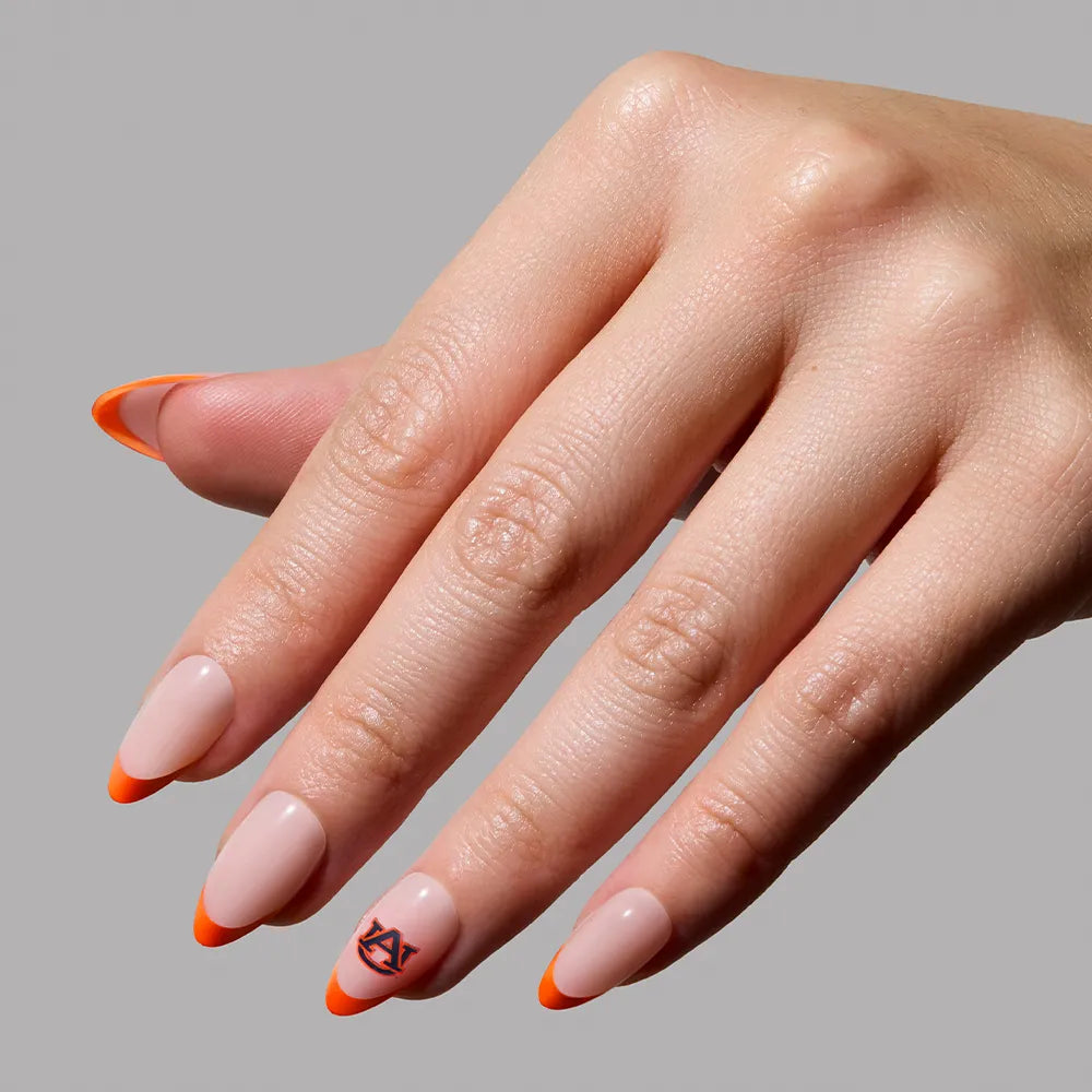 A hand features the glamnetic Auburn Tigers Press-On Nail Set with almond-shaped nails, a nude base, bright orange tips, and a small black geometric design on one tip against a plain gray background. - Product image