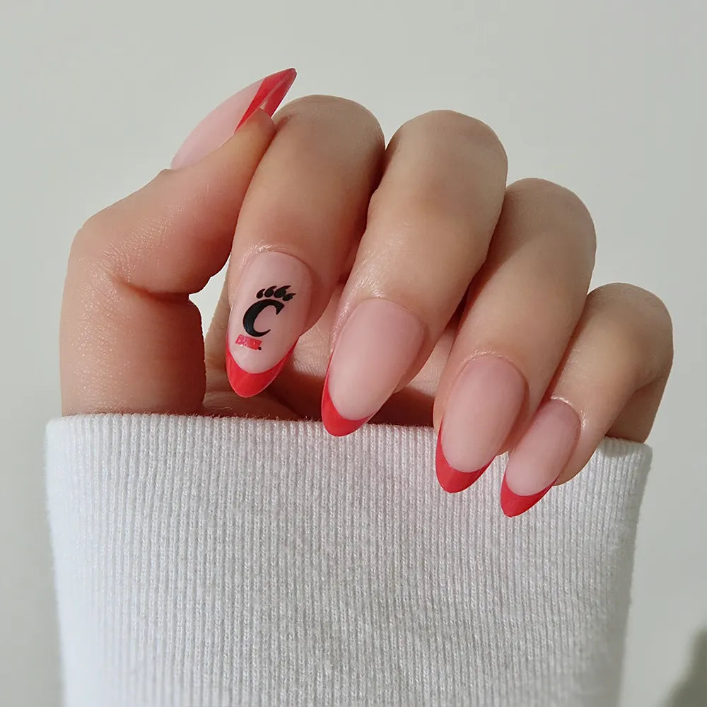 A hand sports glamnetic’s Cincinnati Bearcats Press-On Nail Set: long, almond-shaped nails with a pink base, red tips, and a ring finger featuring a black C and bear paw. The wearer has on a white sleeve.