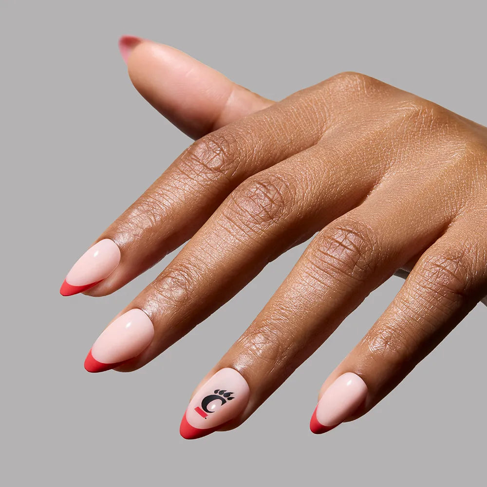 A hand with glamnetic's Cincinnati Bearcats Press-On Nail Set shows almond-shaped, salon-quality nails in light pink with red tips and a black abstract eye graphic on the ring finger against a plain gray background. - Product image