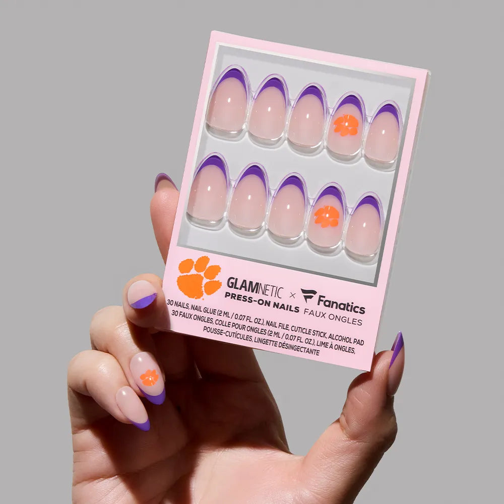 A hand holds the glamnetic Clemson Tigers Press-On Nail Set, featuring clear nails with purple French tips and an orange paw print logo. The pink packaging is accented with bold black and orange text.