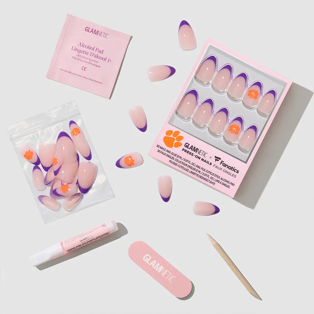 The glamnetic Clemson Tigers Press-On Nail Set includes pink and purple nails with orange paw prints, extra nails, an alcohol pad, nail glue, a nail file, and a cuticle stick arranged neatly on a light surface. - Product image