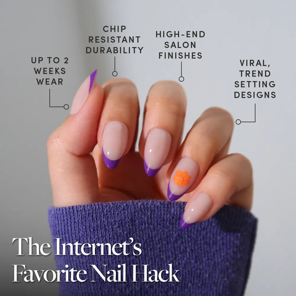 A manicured hand with purple French tip nails and an orange flower shows off the glamnetic Clemson Tigers Press-On Nail Set—durable, trendy, high-end, lasting up to two weeks. Try the Internet’s Favorite Nail Hack. - Product image