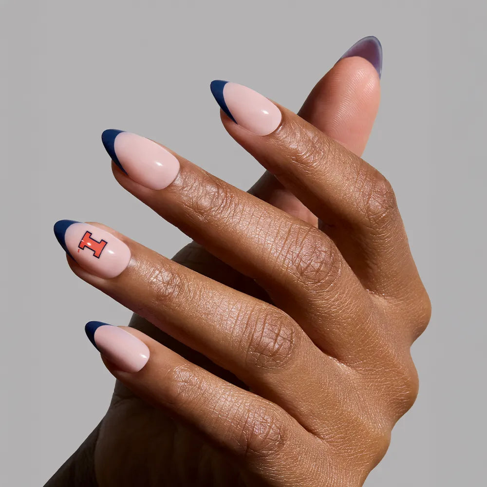 A hand displays glamnetic's Illinois Fighting Illini Press-On Nail Set with almond-shaped, nude nails, blue French tips, and a red and black "I" accent for team spirit. The background is plain gray. - Product image