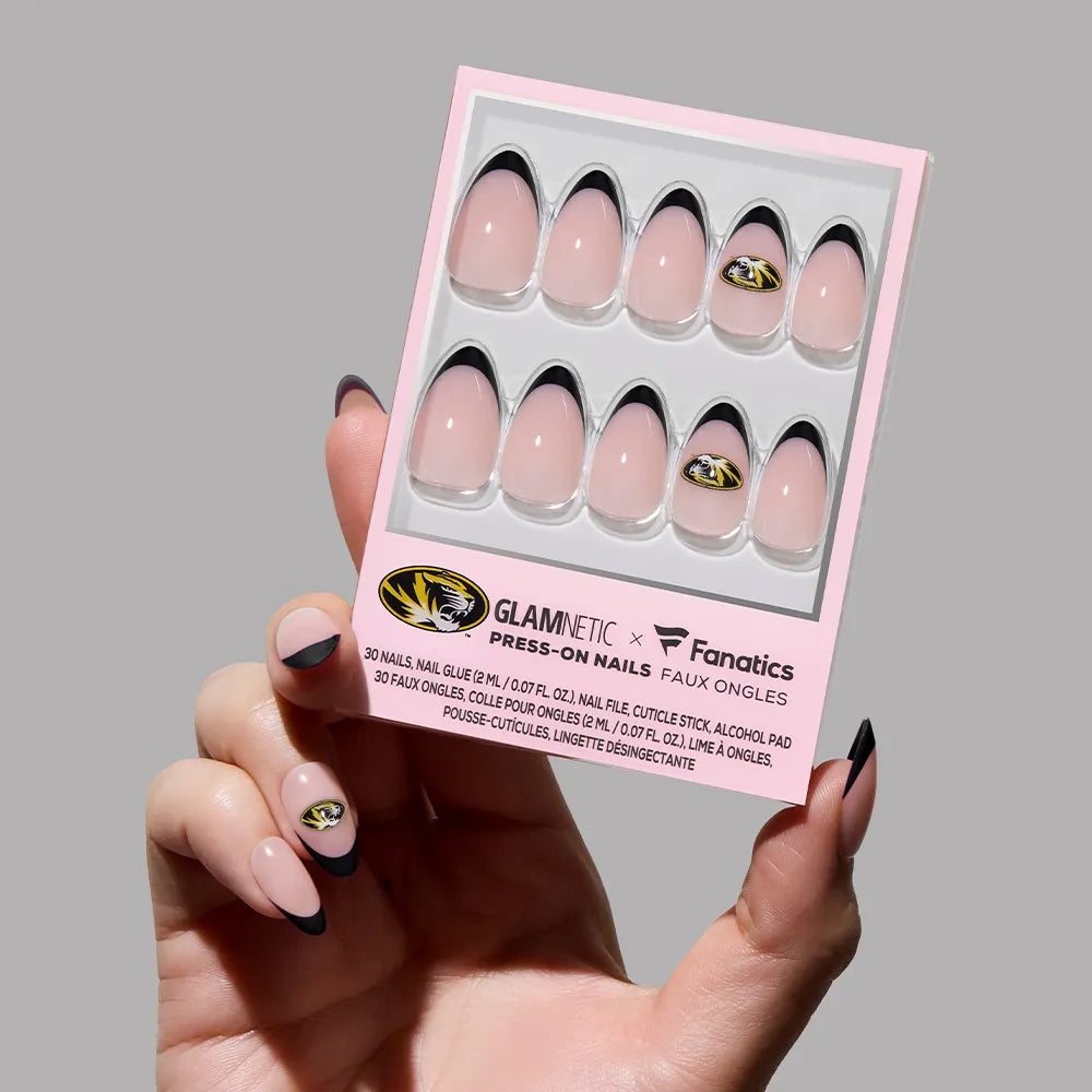 A hand holds a glamnetic Missouri Tigers Press-On Nail Set, showing nude nails with black French tips and an accent nail featuring the team's gold and black logo. - Product image