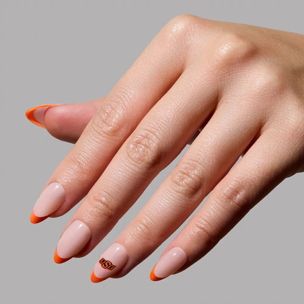 A hand displays the glamnetic Oklahoma State Cowboys Press-On Nail Set with almond-shaped nails, a nude base, bright orange tips, and an orange/black OSU logo on the ring finger. Perfect for fans! Solid light gray background. - Product image