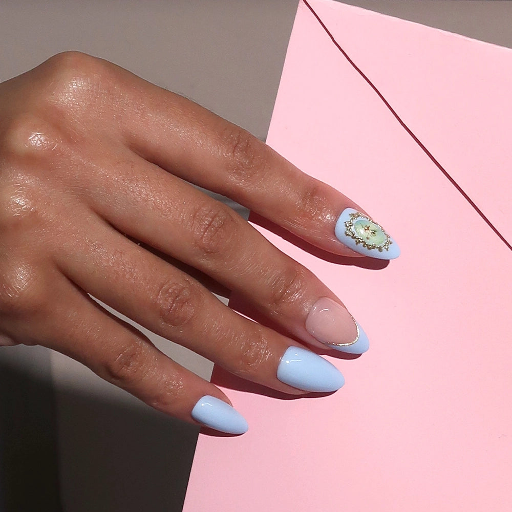 A hand with short almond nails painted in Angel in Blue by glamnetic, featuring light blue and nude tones with a 3D gold gemstone design on the ring finger, rests on a pink envelope. - Product image