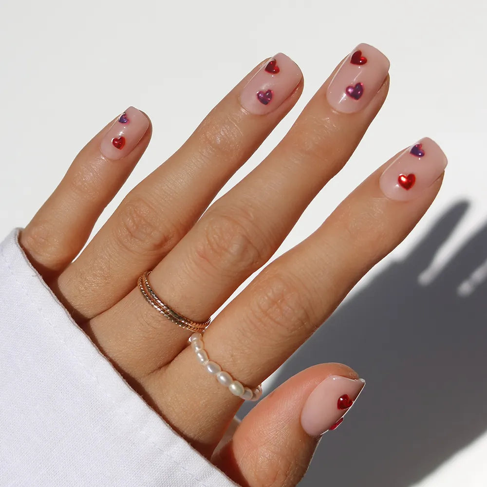 A hand with glossy nude "Dear Lover" nails by glamnetic in a short squoval shape has small red and purple heart accents. Gold and pearl rings adorn the fingers, while a white sleeve partially covers the wrist.