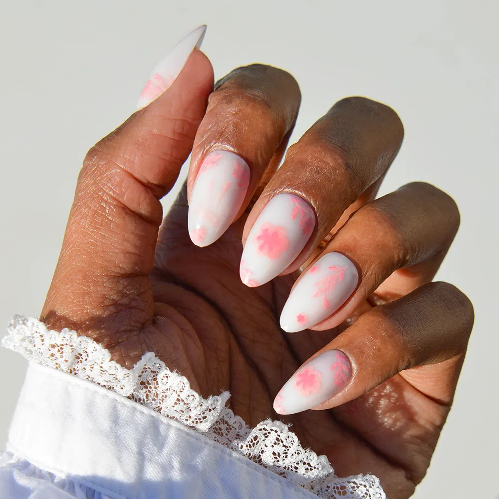 A hand with short almond nails in glamnetic's Fleur Amor style, showcasing a soft white base and delicate pink blossom design. The person wears a white, lace-trimmed sleeve.