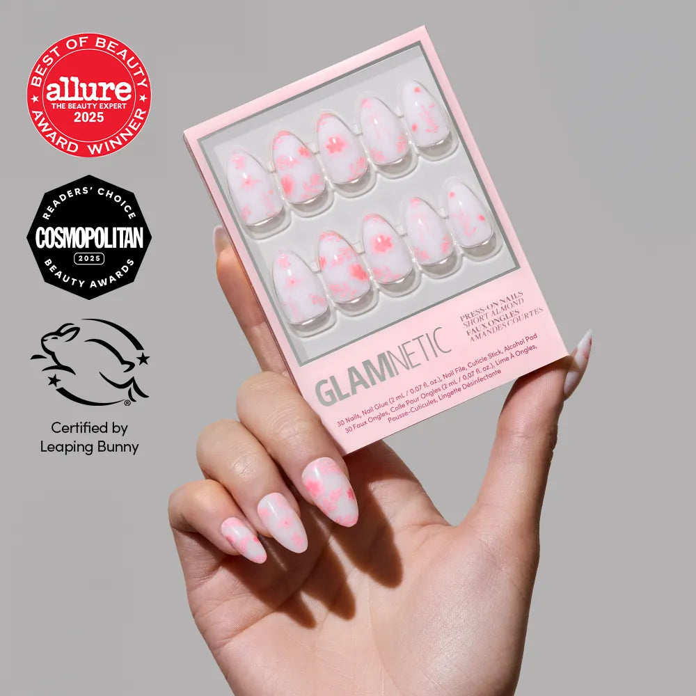 A hand with pink and white cloud-patterned short almond nails holds a matching box of glamnetic Fleur Amor vegan press-on nails. Award logos from Allure, Cosmopolitan, and Leaping Bunny certification are also shown.
