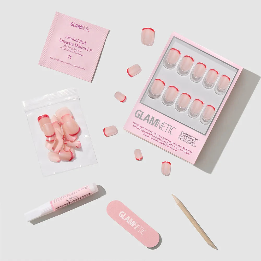 The glamnetic Hotline Red press-on nail set features pink nails with red micro French tips, nail glue, alcohol pad, nail file, cuticle stick, and extra nails—all arranged neatly on a light gray background. - Product image