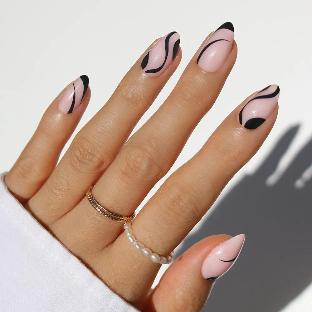A hand with short almond nails painted light pink, featuring abstract black line and swirl designs from the glamnetic Ink Wave set. Gold and pearl rings accent the minimalist look, with a white sleeve partially covering the hand.