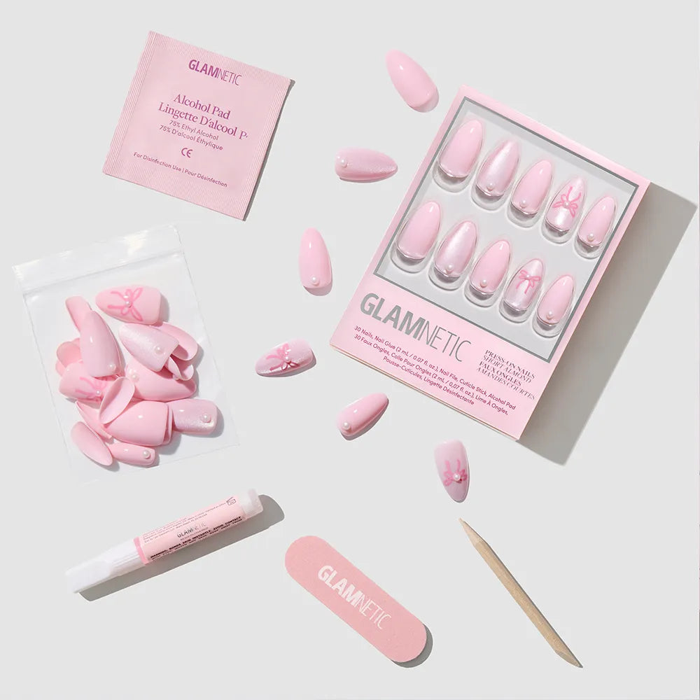 The glamnetic Love Knot set includes pink almond press-on nails with floral designs, a mini file, cuticle stick, adhesive tube, alcohol pad, and extra nails—all for the perfect soft-girl look on a light background. - Product image