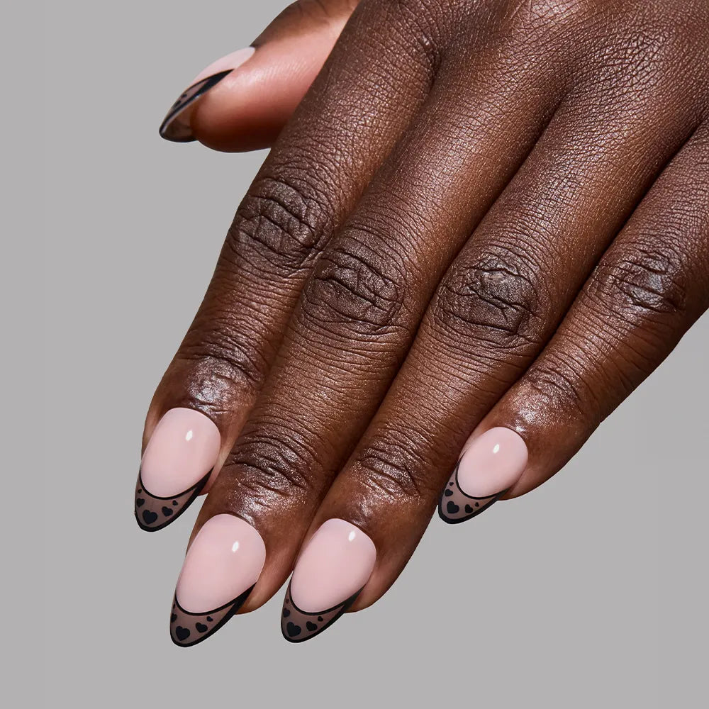 A hand with short almond nails, painted in glamnetic’s Love Noir—a nude shade with black French tips and tiny black hearts—displayed against a plain gray background. - Product image