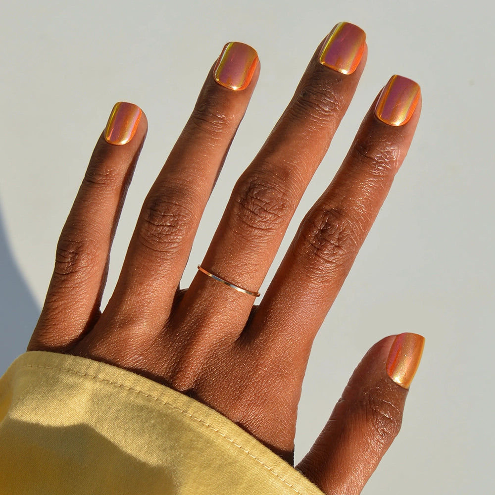 A hand with Papaya Chrome squoval nails by glamnetic, wearing a thin gold ring on the middle finger and partially covered by a pale yellow sleeve, set against a light background.