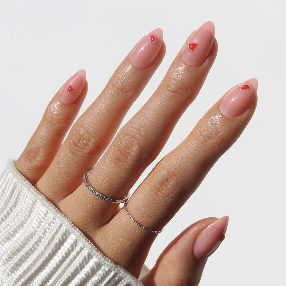 A hand with almond nude pink Juicy Slice Press-On Nails by glamnetic, each tip featuring a small red heart. Two thin silver rings accent the fingers, and a white ribbed sleeve peeks through—ideal for chic, minimal nail lovers.