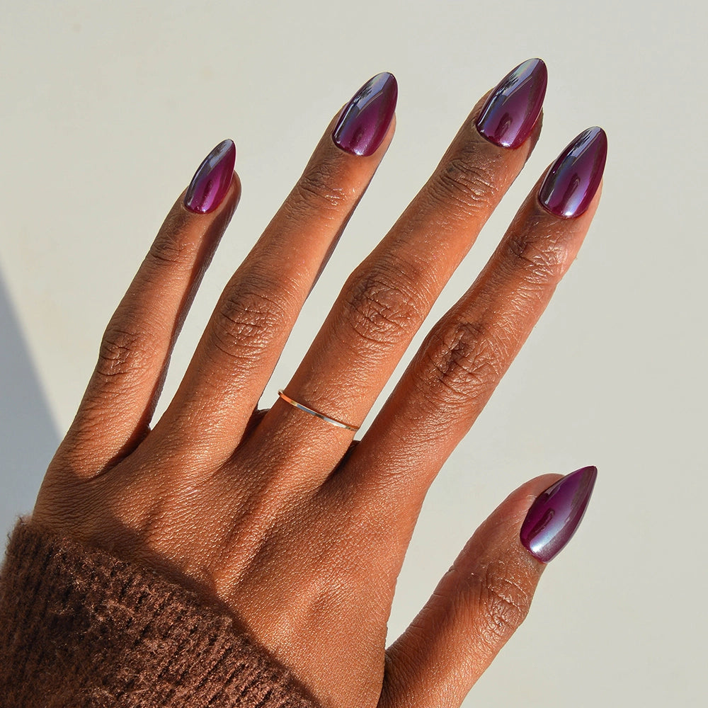 A hand with dark skin shows off long, almond-shaped Jam Drizzle nails by glamnetic in metallic purple. The glossy finish catches the light, paired with a thin gold ring and a brown knit sleeve partially covering the wrist.