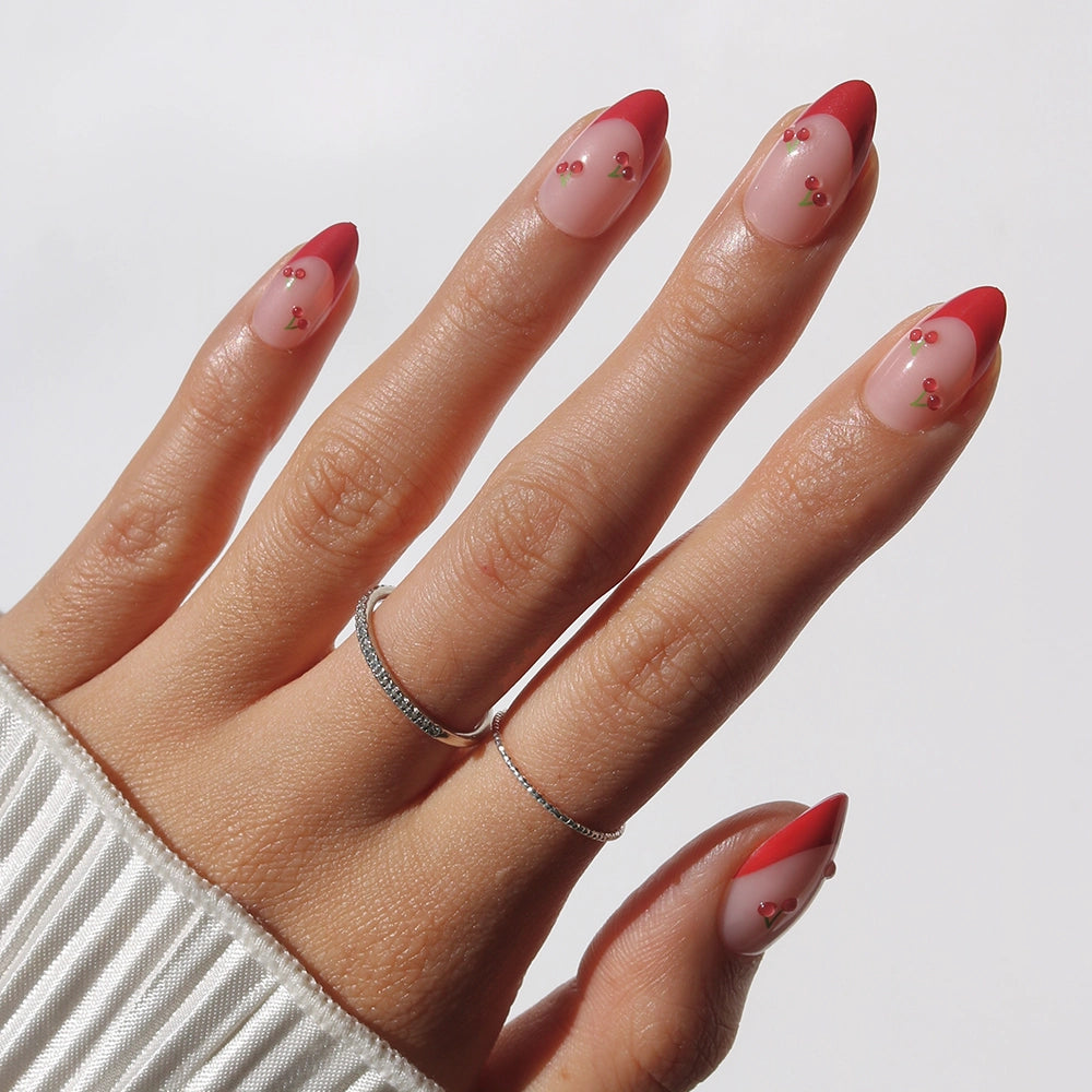A hand with almond nails wears glamnetic's Cherry Bomb red French tips with pink gem accents, creating a chic cherry-inspired look. Two thin silver rings and a white ribbed sleeve complete the style against a plain light background.