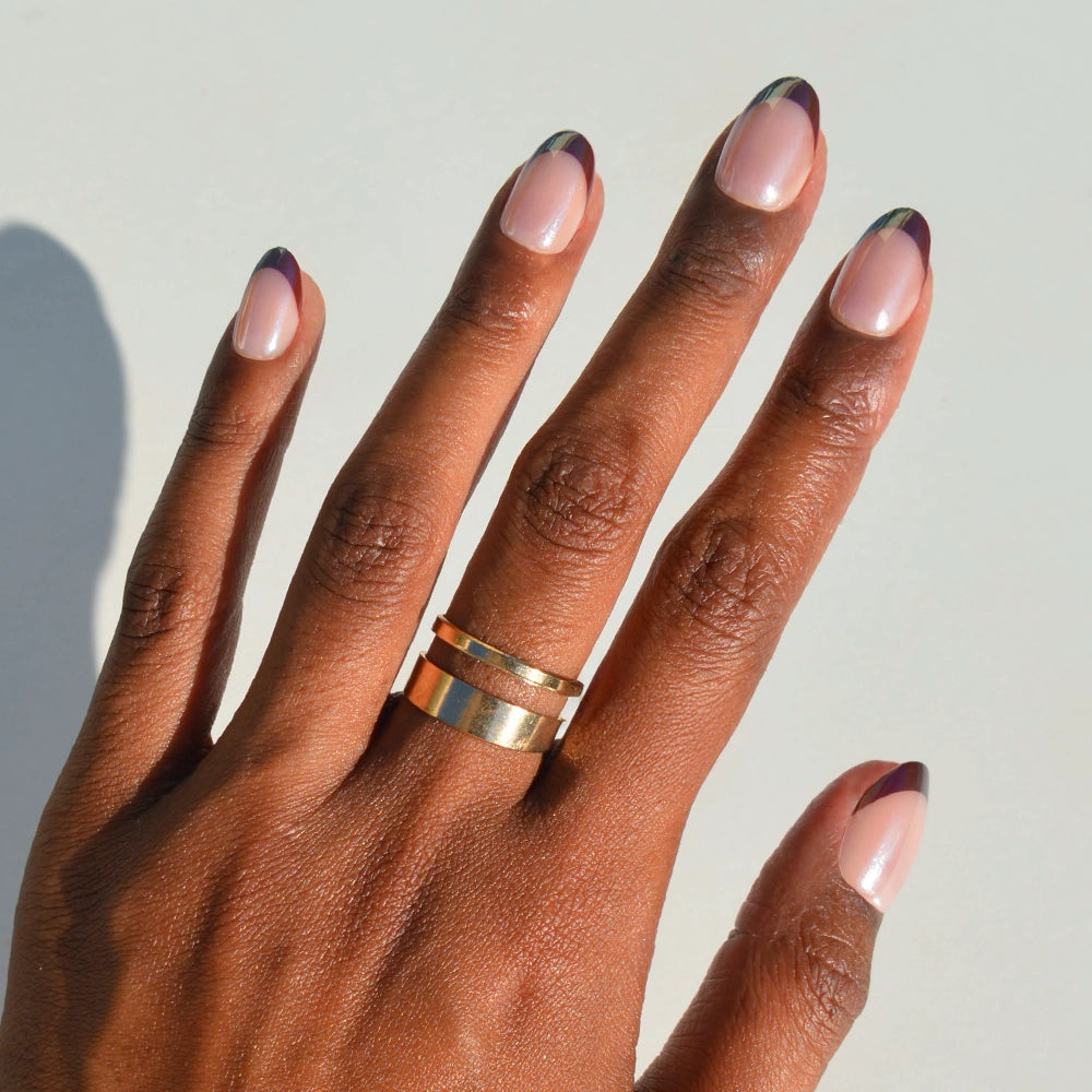 A hand with brown skin wears a gold double-band ring and showcases manicured short oval nails featuring Cherry Mood by glamnetic—chic, glossy press-on nails with a natural base and dark purple and green-tipped French design.