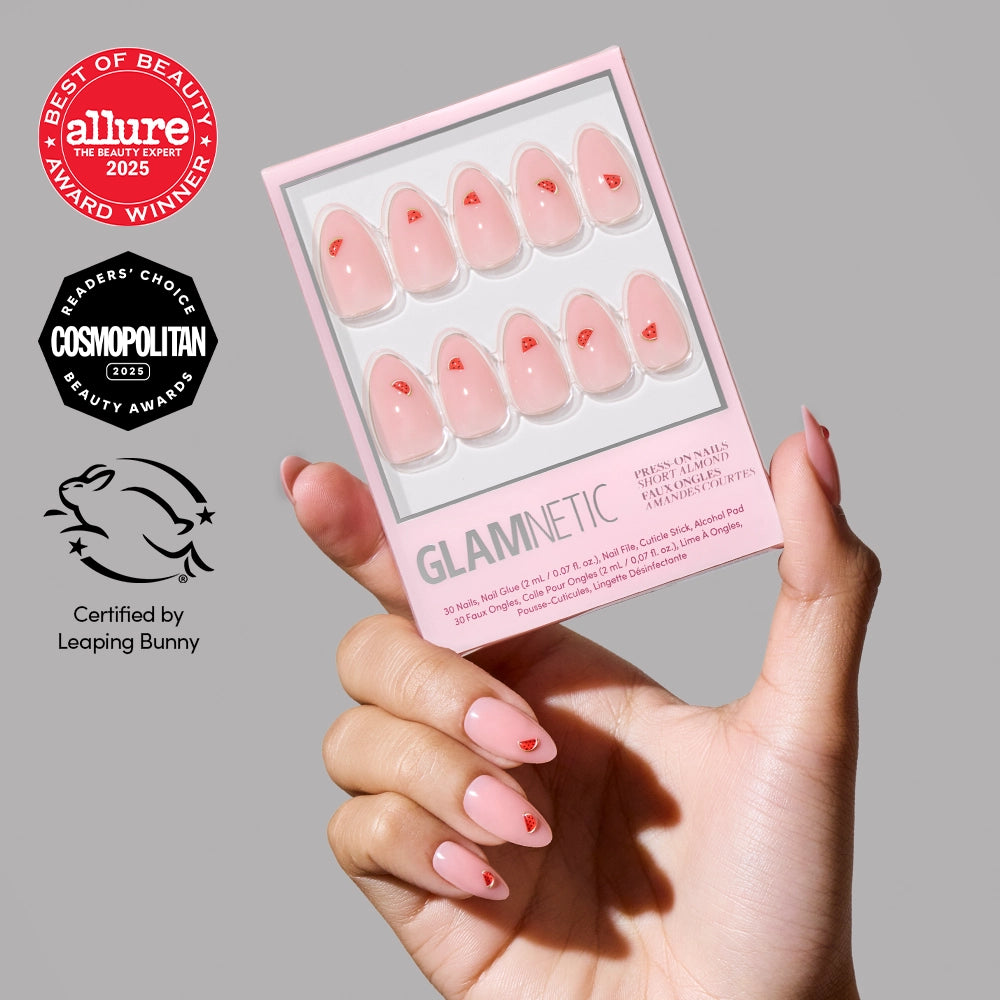 A hand with light pink almond glamnetic Juicy Slice press-on nails, adorned with small red hearts, holds a box of Juicy Slice Press-On Nail sets. Beauty award badges and a cruelty-free certification surround the box.