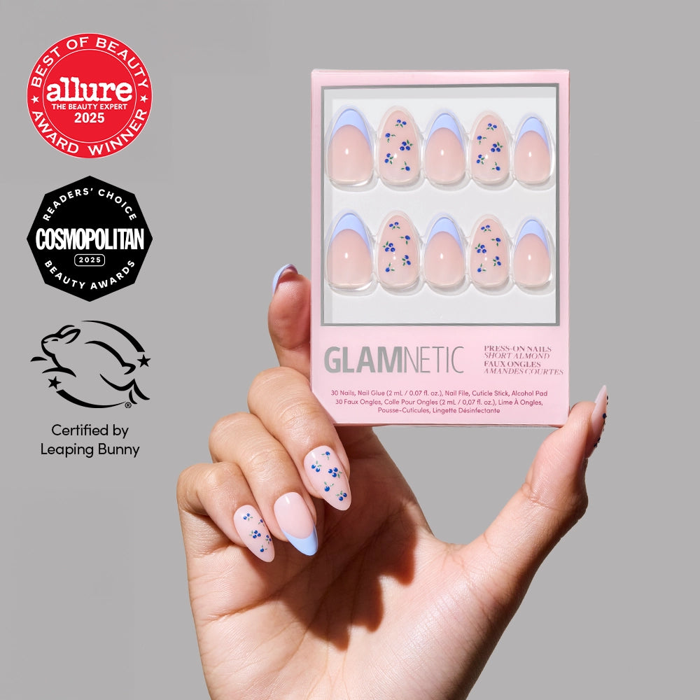 A hand with light pink almond nails featuring blue floral and French tip designs holds a box of glamnetic Bluebelle press-on nails. Allure, Cosmopolitan, and Leaping Bunny certification logos appear on the left.