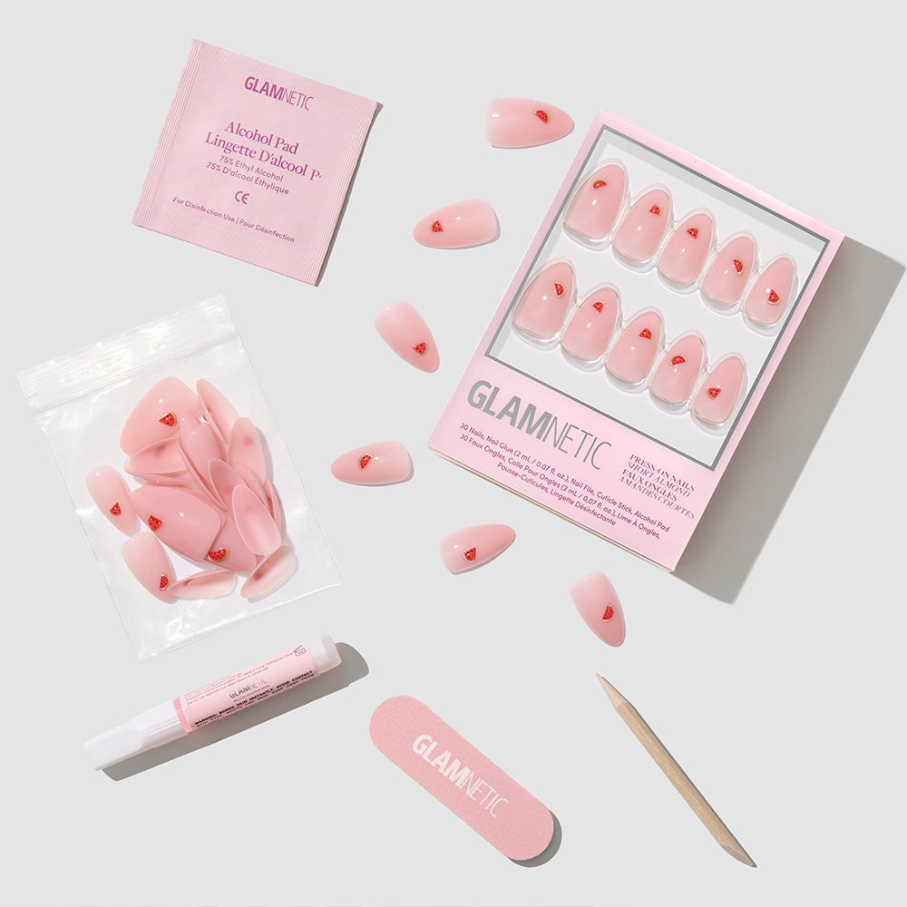 The Juicy Slice Press-On Nails by glamnetic feature pink almond nails with tiny red hearts, plus an alcohol pad, nail glue, file, cuticle stick, and extra nails—all arranged on a light background. - Product image