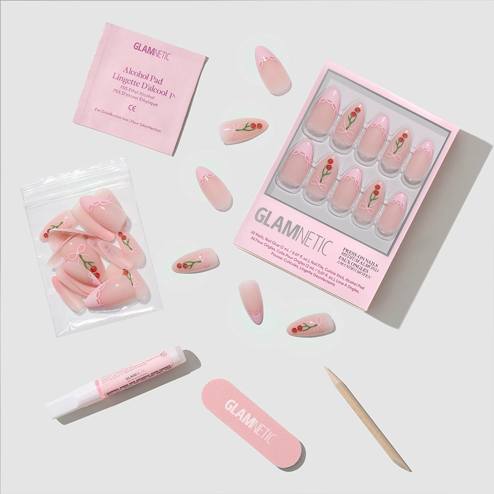 A flat lay of glamnetic Cherry Bow Press-On Nail kit features pink cherry-patterned, almond press-ons, nail glue, a cuticle stick, mini file, alcohol pad, and loose nails arranged on a light background. - Product image