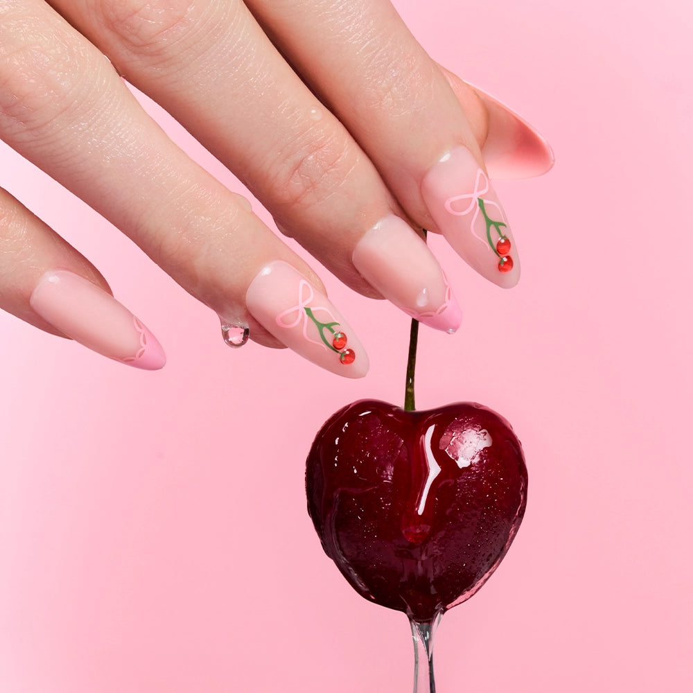 A hand with glamnetic Cherry Bow long, pink French tip nails with cherry and bow designs holds a glossy dark red cherry against a pink background. - Product image