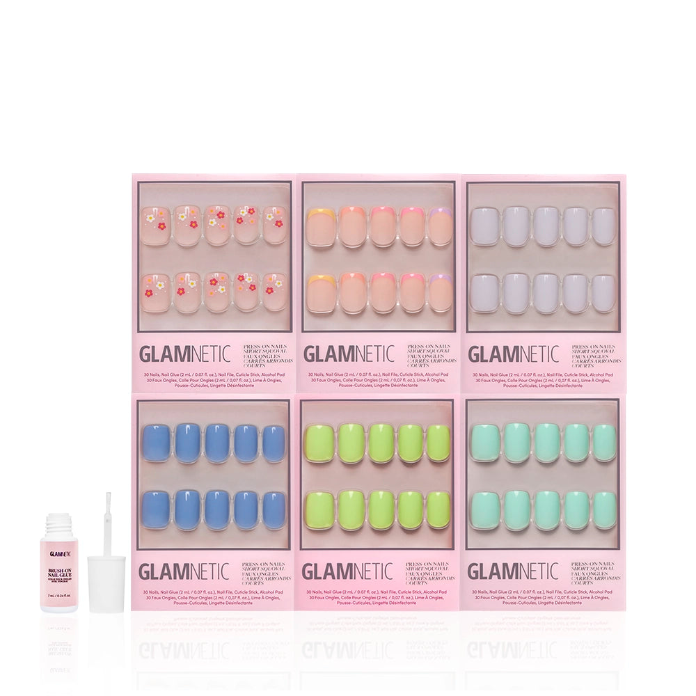 Six sets of glamnetic Squoval Season pastel press-on nails, each with a unique spring-inspired design, are arranged in pink boxes across two rows. A small open bottle of nail glue sits beside the sets on a white background. - Product image