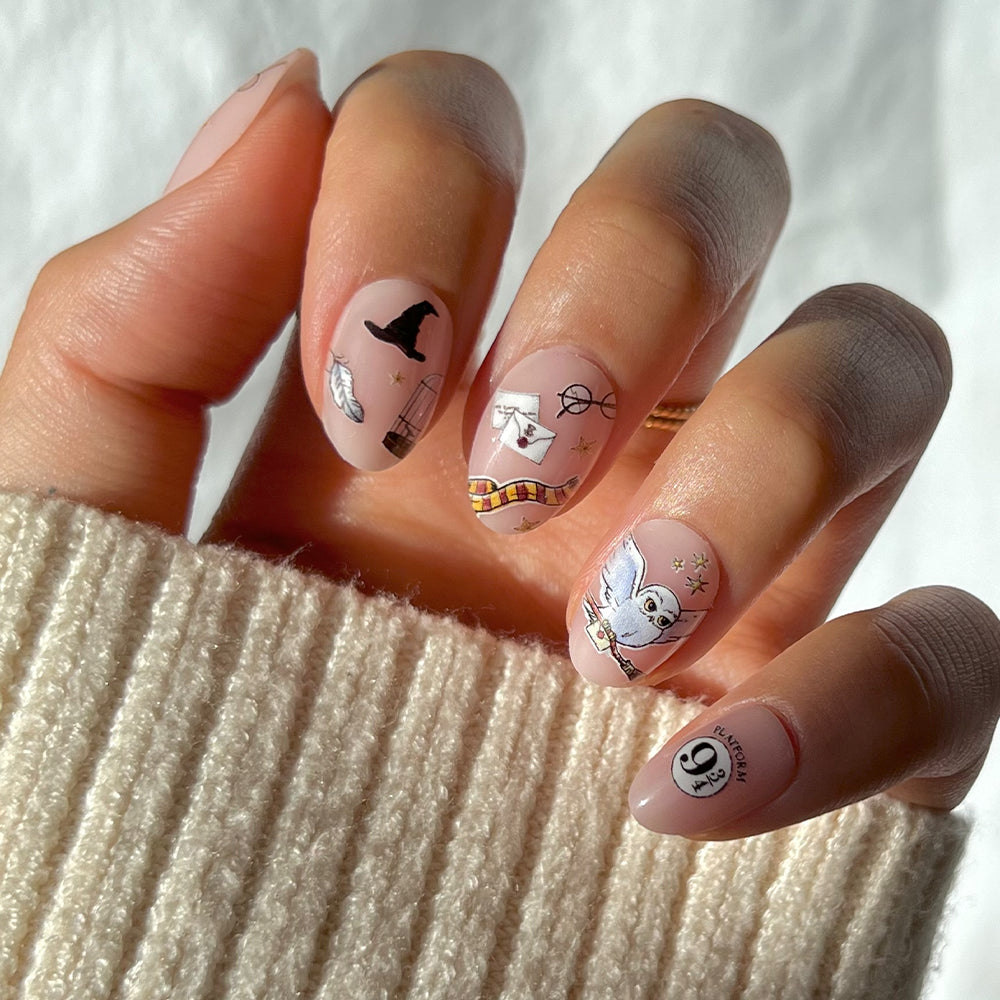 A hand with glamnetic's Hogwarts™ vegan nude press-on nails featuring whimsical Harry Potter designs—witch hat, glasses, Hedwig, scarf, stars, feather, and Platform 9¾—paired with a cozy cream knit sweater.
