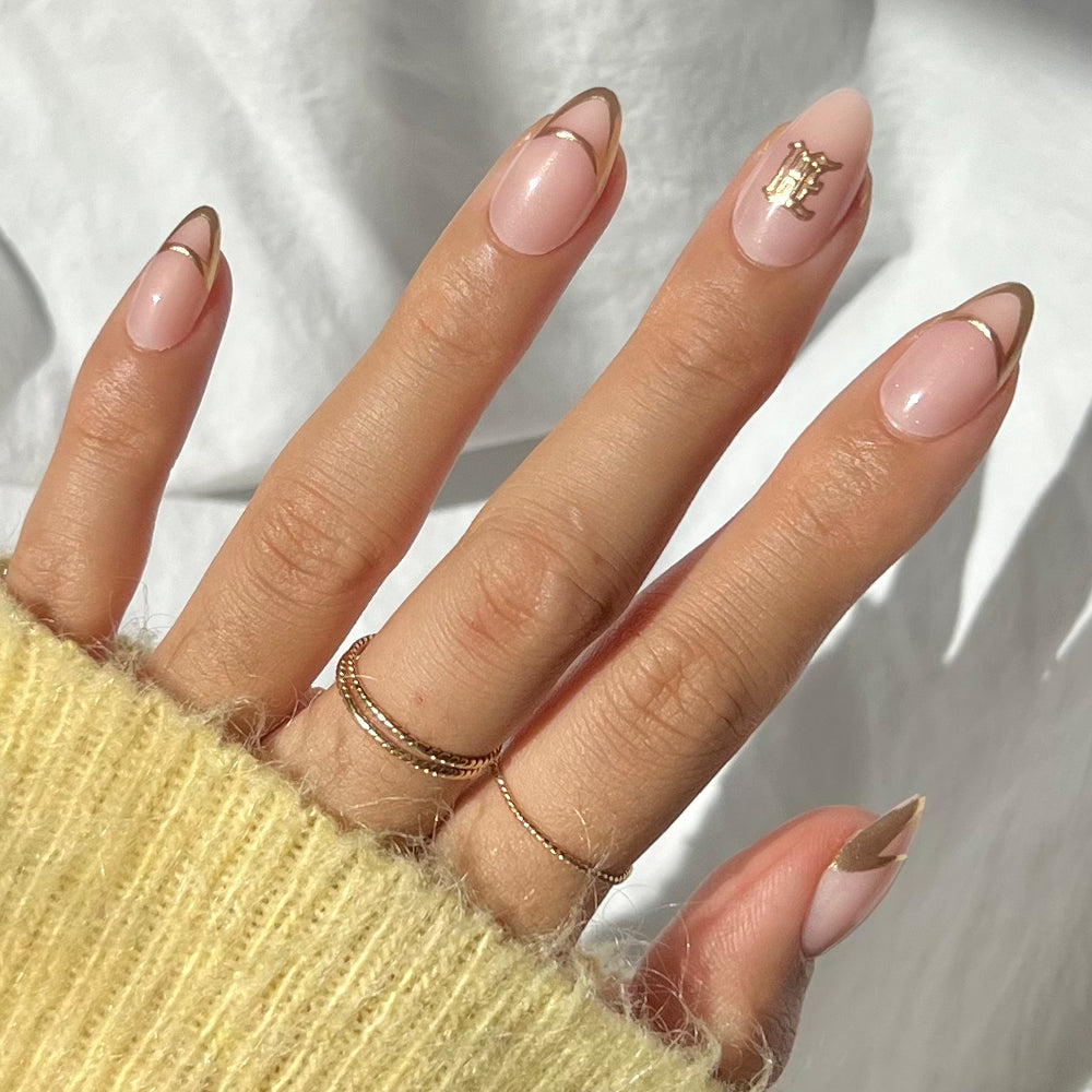 A hand with almond-shaped nails in pale pink and gold French tips showcases the Hufflepuff™ Nail Set by glamnetic. A gold letter accent on the ring finger, delicate rings, and a yellow knit sleeve complete this chic look.