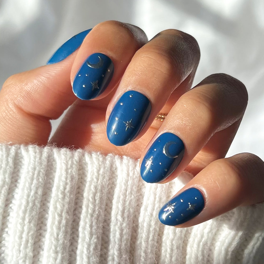A hand with short oval nails painted dark blue, featuring a white moon and star design, wears the Luna Lovegood™ Nail Set by glamnetic. A ribbed white sweater and sunlight highlight the celestial-inspired manicure.