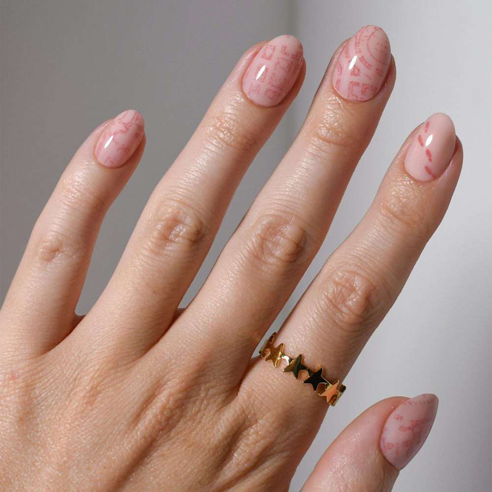 A hand with neatly manicured nails showcases the glamnetic Marauder's Map™ vegan nail set, featuring a pale pink base and light red graphics. The person wears a gold ring with star-shaped cutouts on the middle finger.