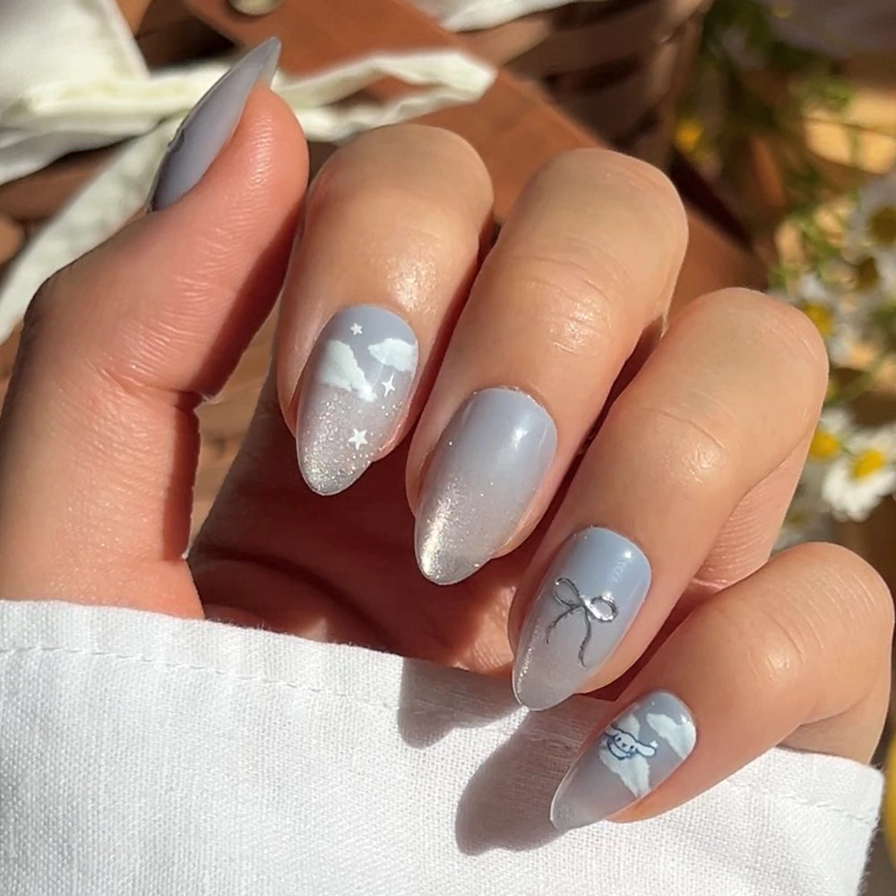 A hand with short almond nails in a light blue and white gradient showcases delicate clouds, stars, a bow, and a bird—designs inspired by glamnetic’s Cinnamoroll™ Cloud set—with a subtle velvet shimmer in sunlight.